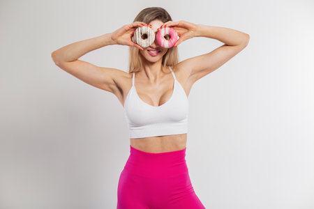 Funny beautiful slim fit woman with smile in fashion sportswear holds with donuts near her eyes on a white background. Sports diet and sweetsの写真素材