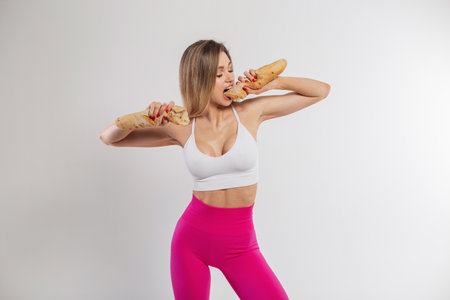 Beautiful fitness girl with a slender body in sportswear holds and bites bread on a white background. Beauty, health, baking and dietの写真素材