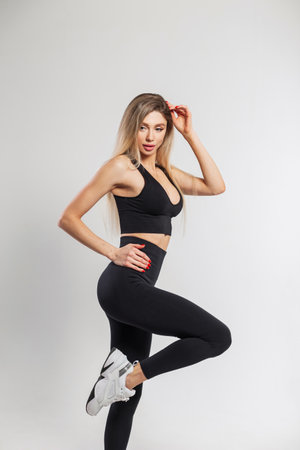 Beautiful athletic fit lady with blond hair in fashionable black sportswear outfit with a top, leggings and sneakers poses on a white background in the studioの写真素材