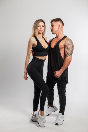 Beautiful athletic young couple in fashionable black sportswear on a white background. Pretty happy fitness girl and handsome strong muscular man are standing in the studioの写真素材