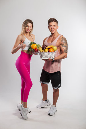 Beautiful slender fitness girl and athletic handsome muscular man holding fruits and vegetables on white background in studio. Healthy vegan foodの写真素材