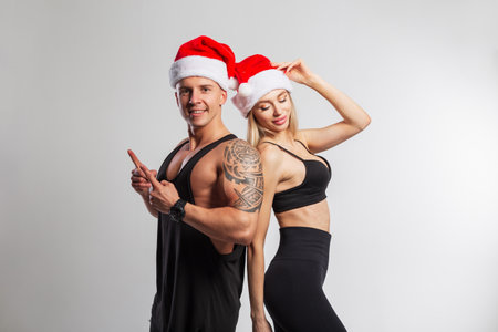 Fashionable beautiful athletic couple in stylish sportswear with a red santa claus hat on a white background. A handsome muscular man and a beautiful fitness lady. New Year and sports lifestyleの写真素材