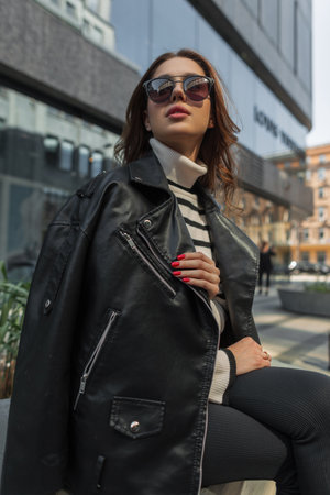 Beautiful fashion young woman model with trendy sunglasses in sweater with black rock leather jacket sits in the city near a modern mallの写真素材