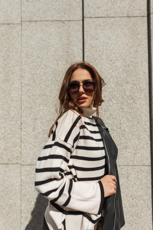 Trendy beautiful fashion young girl with sunglasses in stylish casual clothes with a black leather jacket and a striped sweater in the city near the wallの写真素材