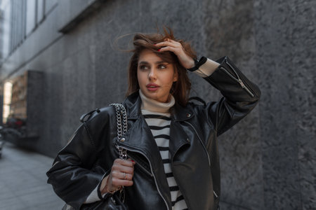 Fashionable beautiful woman with black leather jacket, sweater and bag walks on the street near a...