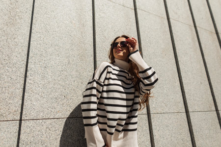 Happy beautiful stylish woman in a trendy fashion striped sweater stands near the wall and puts on sunglasses on a sunny day. Urban female style look fashionの写真素材