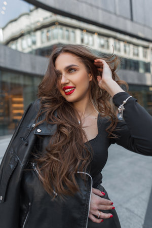 Happy beautiful fashionable fashion chic woman with red lips with a smile in fashion black clothes with a stylish leather jacket walks in the city. urban party girlの写真素材