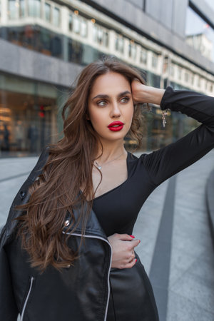 Street female fashion portrait of a beautiful stylish chic lady with red lips in trendy black clothes with a leather jacket posing in the cityの写真素材