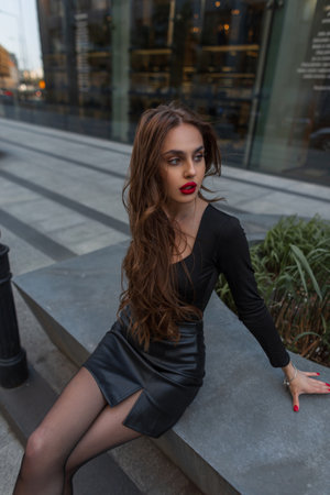 Beautiful fashionable chic lady in stylish elegant clothes with red lips with black top and black skirt sits in the cityの写真素材