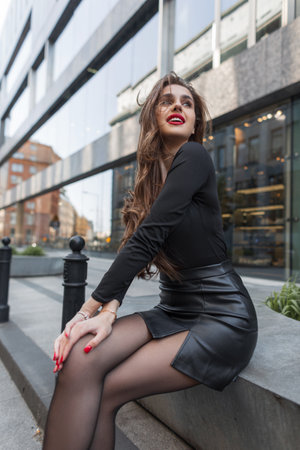 Happiness fashion pretty chic woman with smile with red lips in black stylish elegant clothes with leather skirt sits on the streetの写真素材