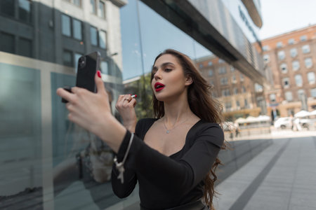 Beautiful fashionable chic lady holds a phone and paints her lips with red lipstick, walks in the cityの写真素材