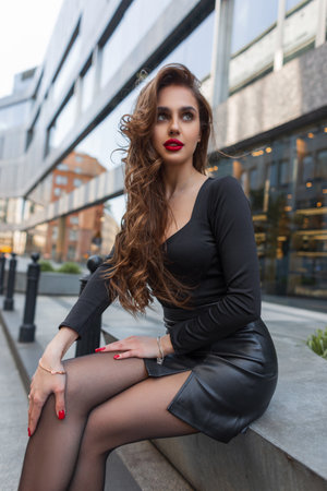 Beautiful fashionable stylish woman with red lips and sexy slender legs in a trendy fashion leather skirt is sitting in the city near a modern buildingの写真素材