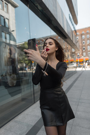 Beautiful young pretty chic woman model paints her lips with red lipstick and looks into a smartphone in the cityの写真素材