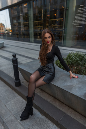 Beautiful fashionable sexy woman with red lips with slender legs in fashion elegant clothes with a skirt, tights and heels is sitting in the cityの写真素材