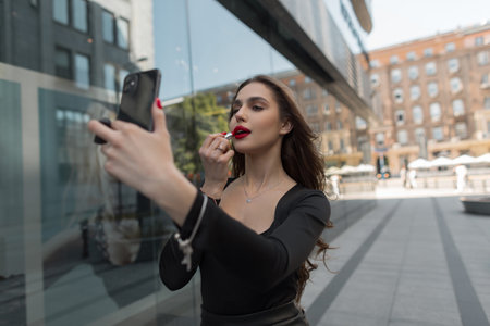 Fashion beautiful chic girl in black elegant clothes walks in the city looks into a smartphone and does makeup red lips. party womanの写真素材