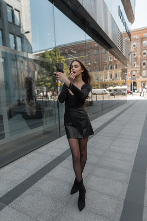 Beautiful sexy fashionable chic woman with slender legs in elegant clothes with a skirt paints her lips with red lipstick and looks into a smartphone on the streetの写真素材