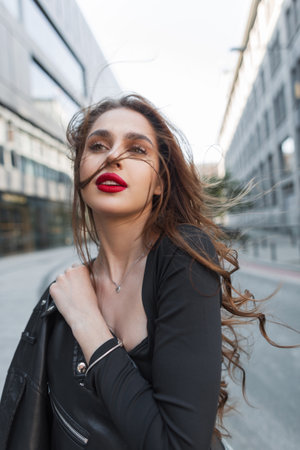 Beautiful elegant chic girl with red lips in fashion black clothes with a leather jacket walks in the city on a sunny dayの写真素材