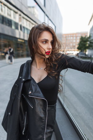 Elegance fashionable young beautiful chic woman with red lips in black fashion clothes with leather jacket walks in the cityの写真素材