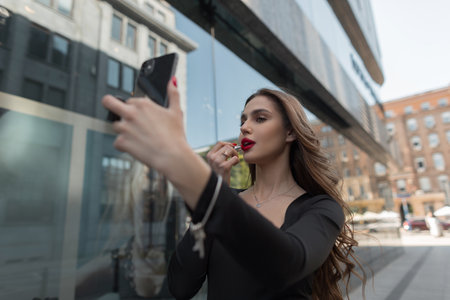 Gorgeous beautiful young fashion chic girl model paints her lips with red lipstick and looks into a smartphone in the cityの写真素材