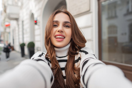 Funny beautiful young woman with a white cute smile in a trendy fashion striped sweater travels and takes a selfie photo in the city near a vintage buildingの写真素材