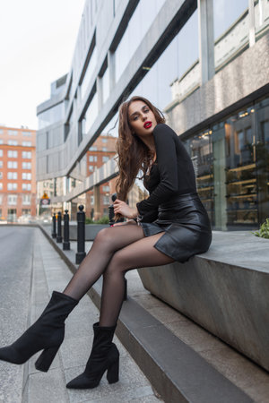 Beautiful fashionable sexy woman with red lips with slender legs in a fashion leather skirt with tights and heels sit and pose in the cityの写真素材
