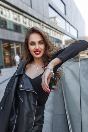 Happy beautiful young fashionable girl with red lips and a smile in black elegant clothes with a leather black jacket walks and poses in the cityの写真素材