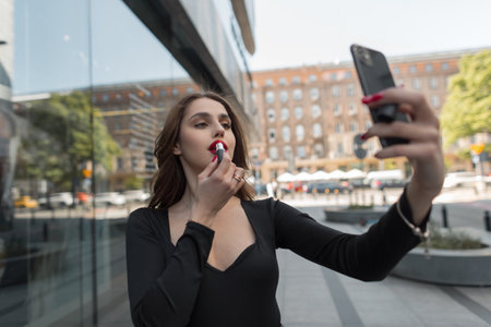 Beautiful young fashion woman model in stylish elegant clothes walks in the city, holds a smartphone and paints her lips with red lipstickの写真素材