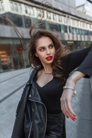 Fashionable beautiful chic girl with red lips in black stylish clothes with leather jacket walks in the city on a windy dayの写真素材