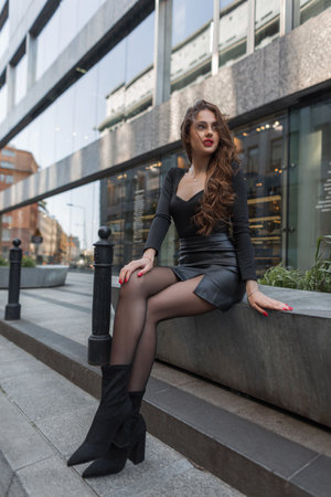 Beautiful stylish young gorgeous chic woman with red lipstick with slender sexy legs in a fashion leather skirt sits in the city near the mallの写真素材