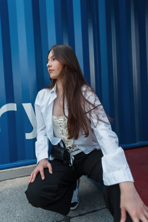 Stylish beautiful fashion youth girl in fashionable clothes with a white shirt sits near a blue metal wall on the streetの写真素材