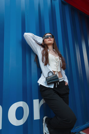 Young beautiful stylish girl in fashionable clothes stands and poses near the blue metal wall on the streetの写真素材