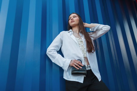 Stylish young woman model in fashion clothes with a white shirt, a corset and a handbag stands near a blue metal wall on the streetの写真素材