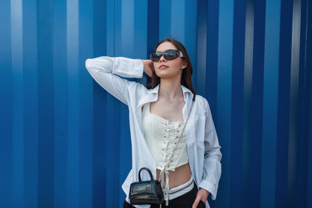 Fashionable beautiful young teenager girl with sunglasses in stylish clothes with a shirt, corset and handbag stands near a blue metal wallの写真素材