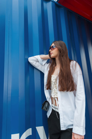 beautiful youth girl in stylish clothes with sunglasses and a white shirt with a handbag stands near a blue metal wallの写真素材