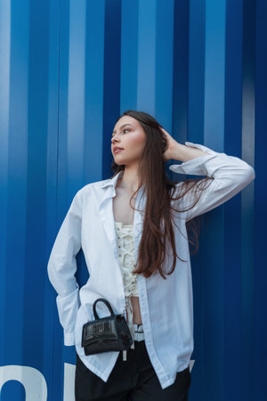 Beautiful young fashionable woman model in stylish youth clothes with a white shirt and a handbag stands near a blue metal wallの写真素材