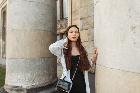Fashionable beautiful young woman in stylish clothes with a white shirt and black dress with a handbag near a vintage building with columns in the cityの写真素材