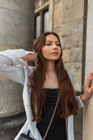 Fashionable beautiful stylish chic girl in trendy fashion clothes with a white shirt and a black dress with a bag walks in the city near the vintage building columnの写真素材