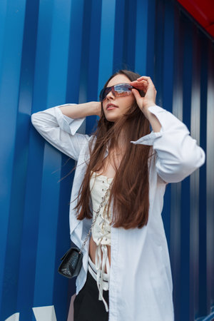 Fashionable beautiful young girl in trendy fashion clothes with bag puts on stylish sunglasses and stands near the blue metal wallの写真素材