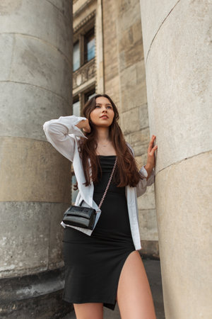 Stylish beautiful young woman model with handbag in a white fashionable shirt and black dress stands near the column in the cityの写真素材