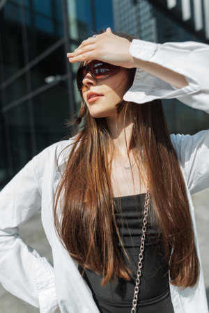 Cool trendy pretty chic youth girl with stylish sunglasses in trendy fashion summer outfit covers her eyes with hand and walks in the city on a sunny dayの写真素材