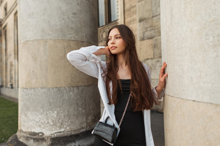 Fashionable beautiful young caucasian girl model with bag in a stylish black dress with a white shirt walks near a vintage building with columnsの写真素材