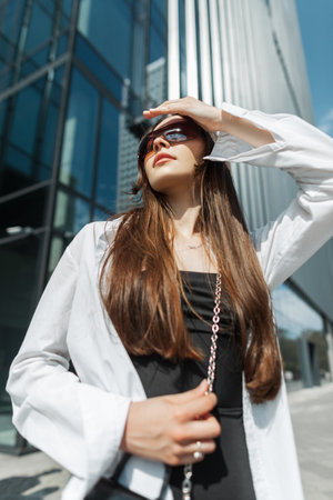 fashionable beautiful young woman with sunglasses in stylish clothes with a black dress and a white shirt with a handbag walks in the city on a sunny day and covers her eyes with handの写真素材