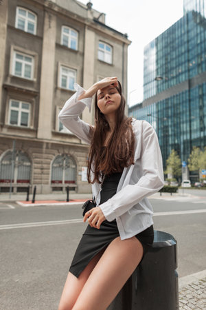 Beautiful fashion elegant chic sexy woman in a stylish black dress with a white shirt and a bag sits in the city and straightens her hairの写真素材