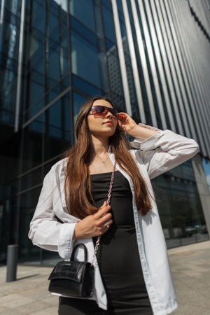 Fashionable elegant pretty lady in trendy fashion clothes with black dress, white shirt with a handbag walks in a sunny modern city and puts on sunglassesの写真素材