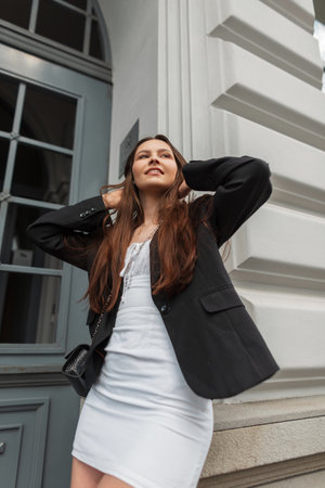 Fashion beautiful happy woman with smile in stylish casual outfit with white dress, black blazer and handbag stands near a vintage building on the streetの写真素材