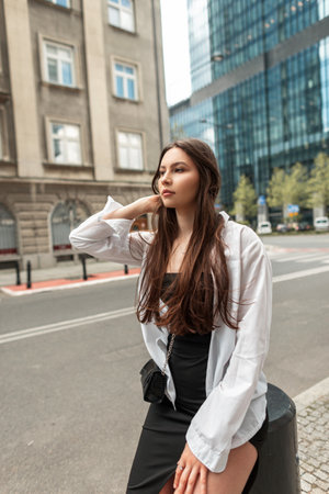 Stylish beautiful young chic woman model with brunet hair in fashion clothes with a black dress and a white shirt with a bag sits in the cityの写真素材