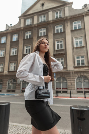 Fashionable beautiful stylish girl in stylish clothes in a black dress with a white shirt and a black handbag walks on the street in the cityの写真素材