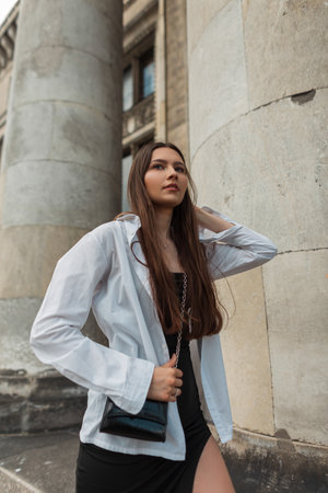beautiful young elegant chic girl in stylish clothes with a white fashion shirt and a black dress with a handbag walks near a vintage building with columns in the cityの写真素材