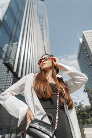 Beautiful young stylish fashionable girl with sunglasses in fashion trendy clothes with a stylish black dress with a white shirt and a handbag walks in the city near a modern business buildingの写真素材