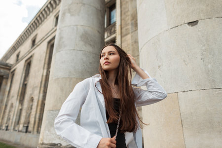 Fashionable beautiful street girl model in stylish casual clothes with a white shirt and black dress with a bag is walking near a vintage building with columnsの写真素材
